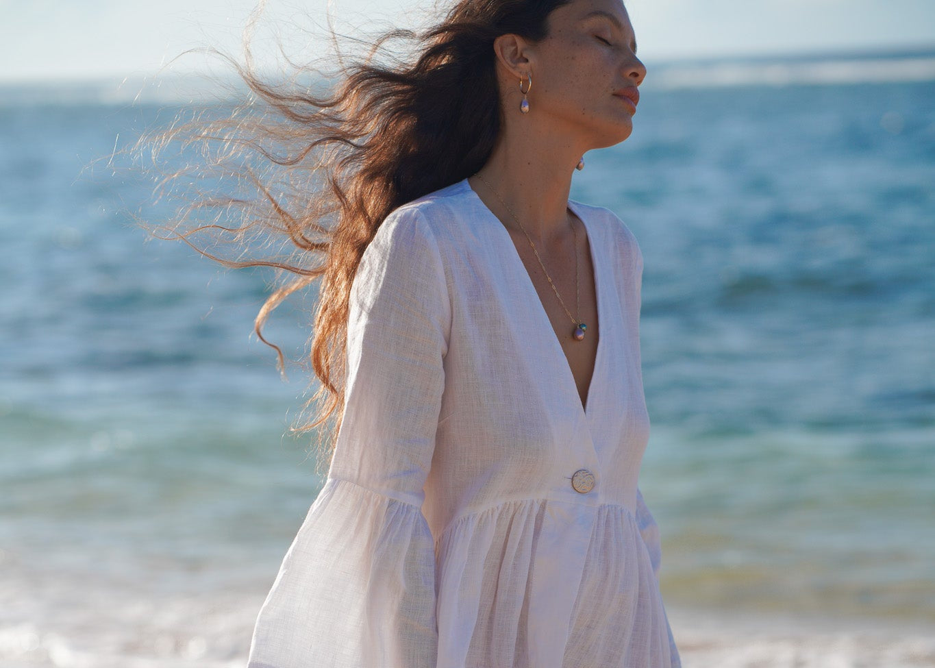 Woman in a white linen dress standing on a beach with ocean in the background