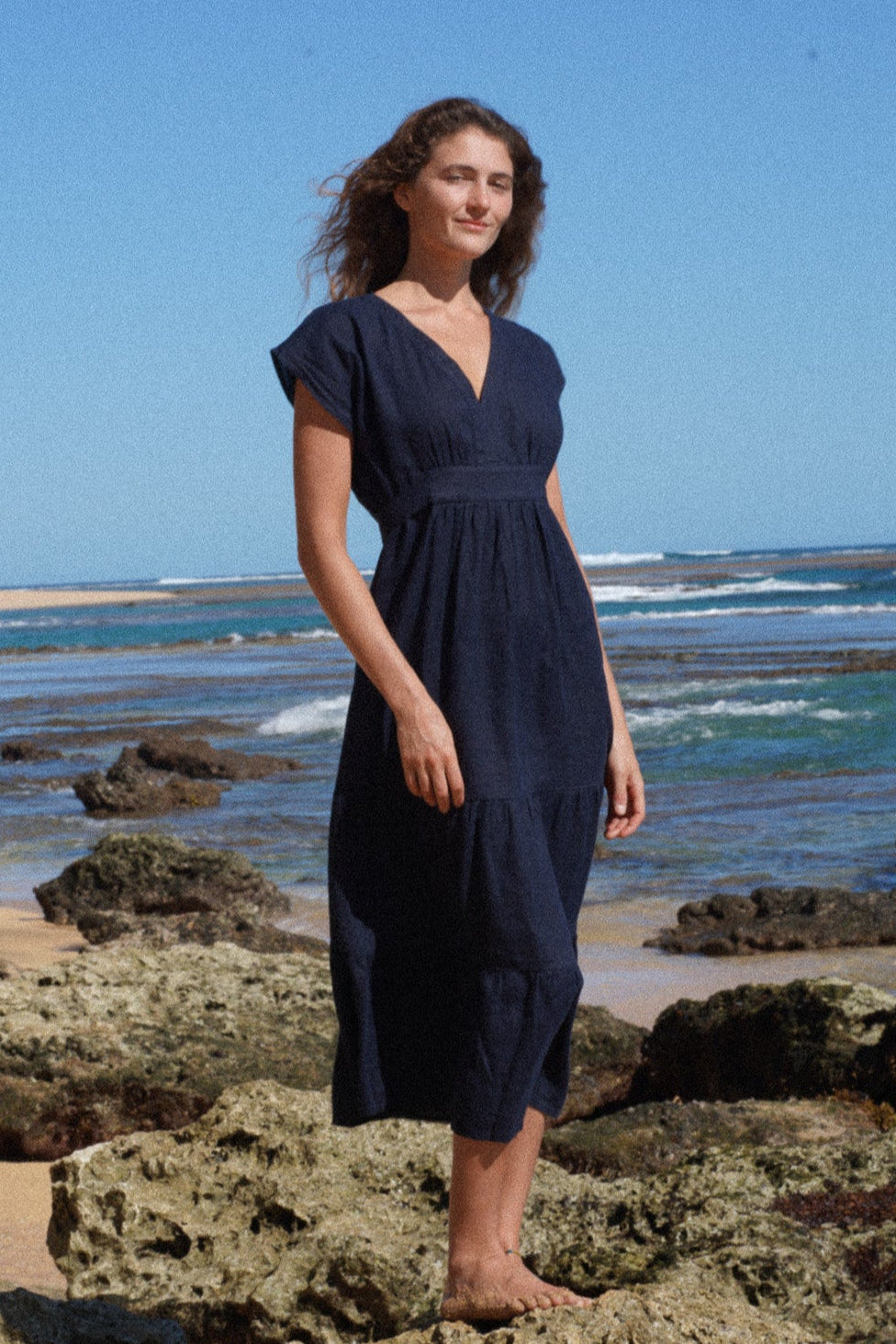 Woman in a navy blue french linen dress standing on rocks by the ocean