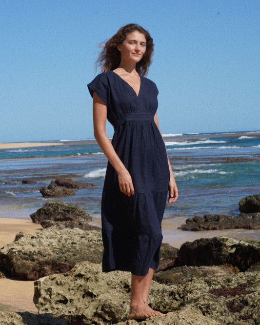 Woman in a navy blue french linen dress standing on rocks by the ocean