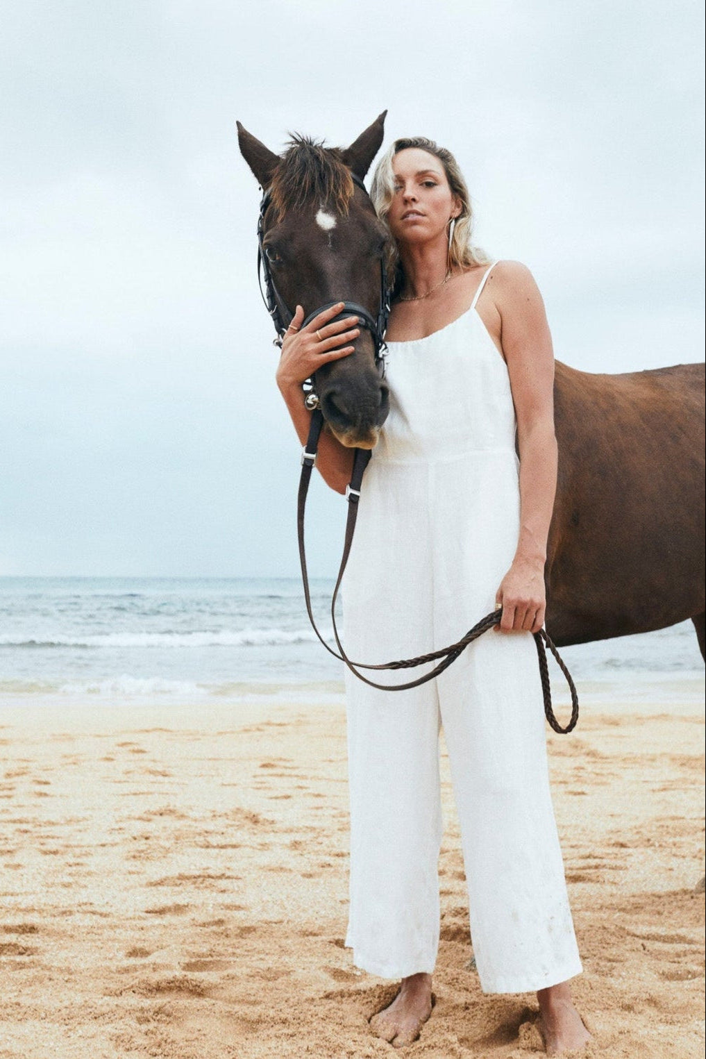 A woman in the white Isabella linen jumpsuit holds a horse close on the beach