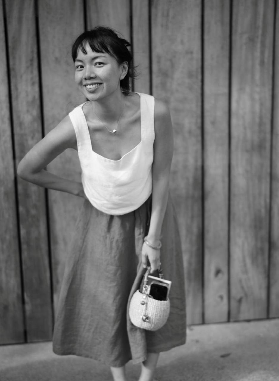 Isabella in a white linen top and linen skirt, smiling, a woven basket bag in hand