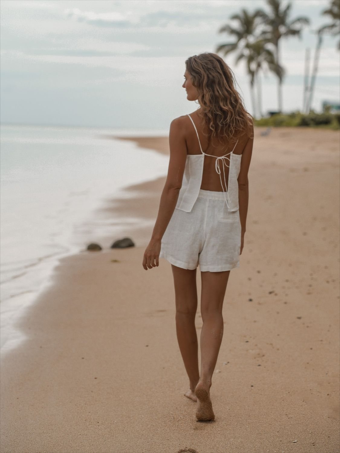 The white Bella Drawstring Top, open at the back, with linen shorts on the shoreline